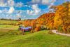 Rustic red farm structures in rolling green hills under a bright cloudy sky are surrounded by brightly colored red and yellow leafed trees.