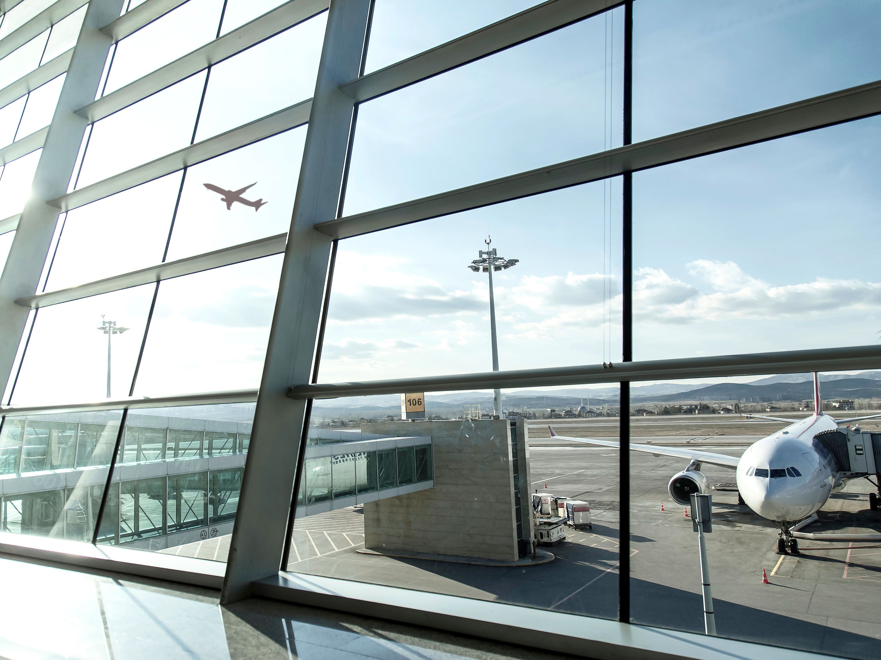 Looking out a window from an airport gate area to a plane taking off and another plane attached to a jetway.