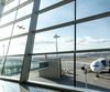 Looking out a window from an airport gate area to a plane taking off and another plane attached to a jetway.