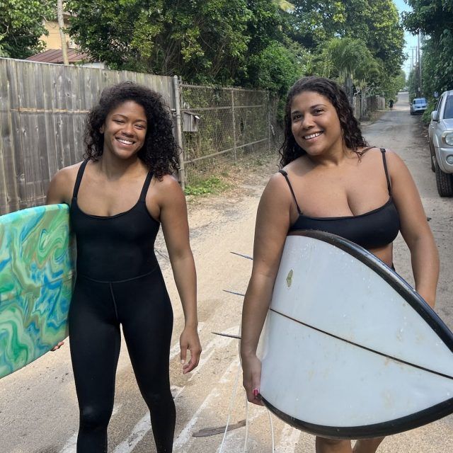 Surfers Chelsea Woody and Tara Crystal walk down the road holding their surfboards and smiling