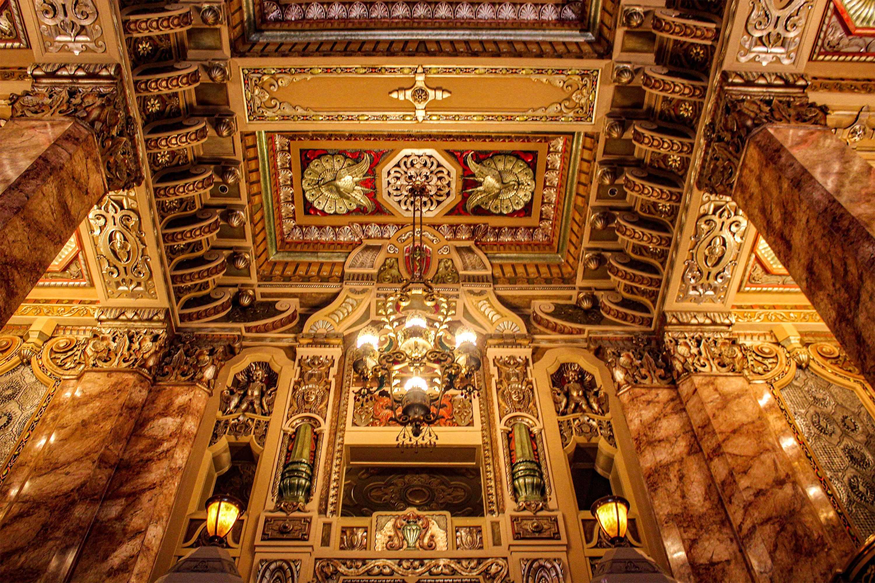 A close-up view of a lavishly decorated theater ceiling with detailed carvings, gold accents, and a hanging antique chandelier.