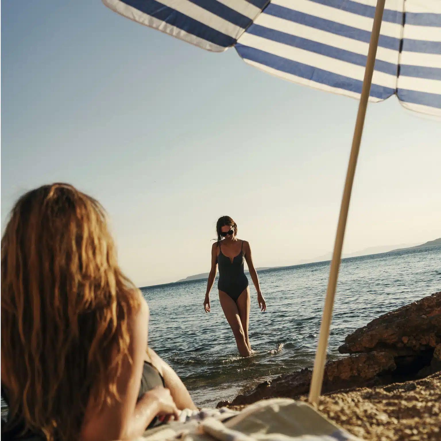 A woman in a black swimsuit and sunglasses walks out of the sea towards a friend sunba a stripy beach umbrella 