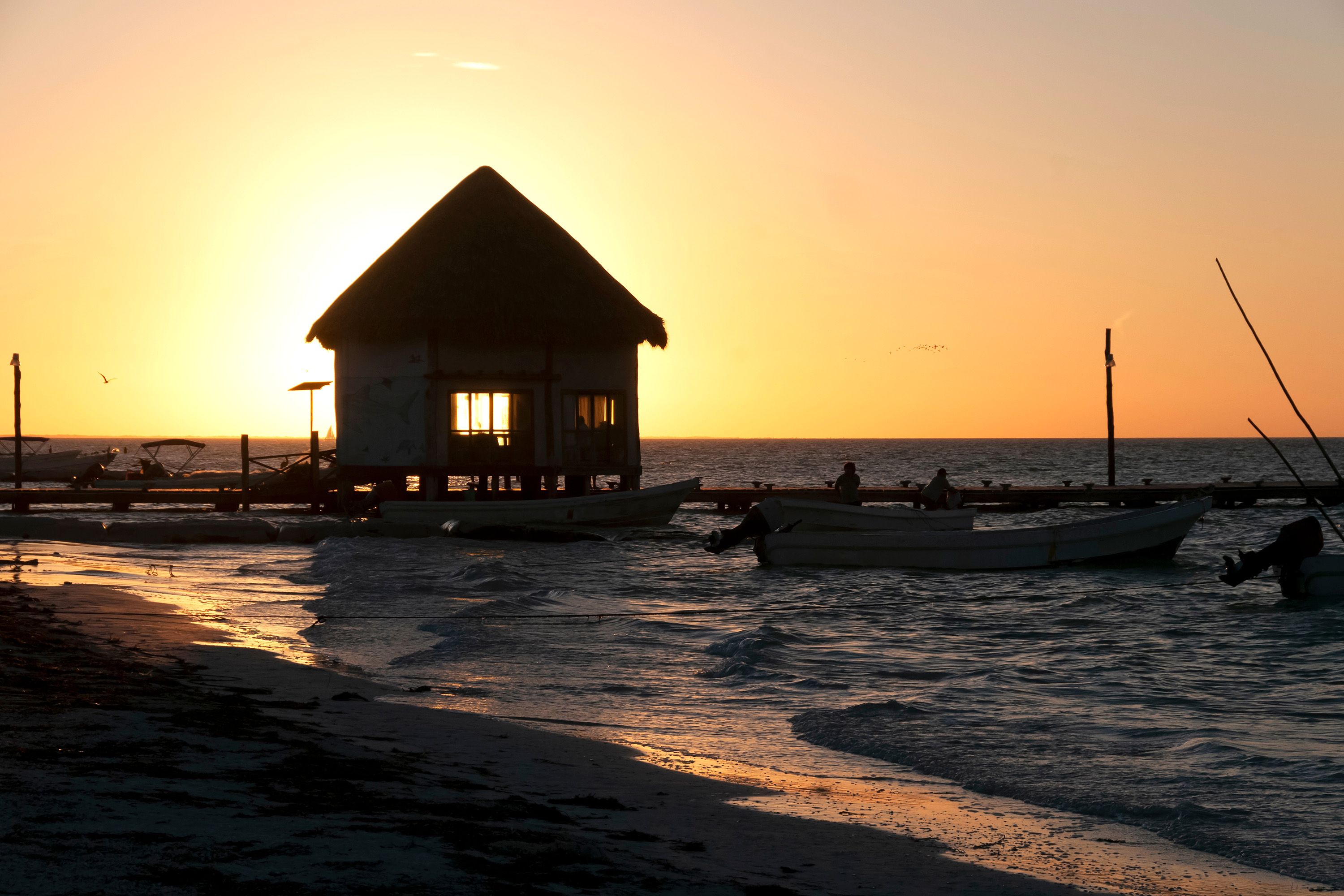 The sun sets over a beach shack with a dock and boats.