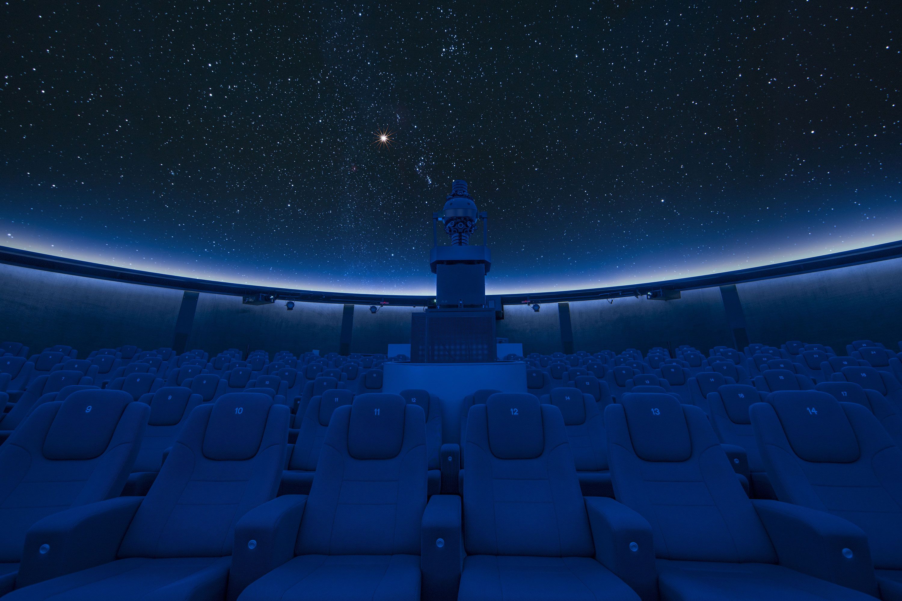 Interior view of darkened planetarium with seating and projector and stars on ceiling.