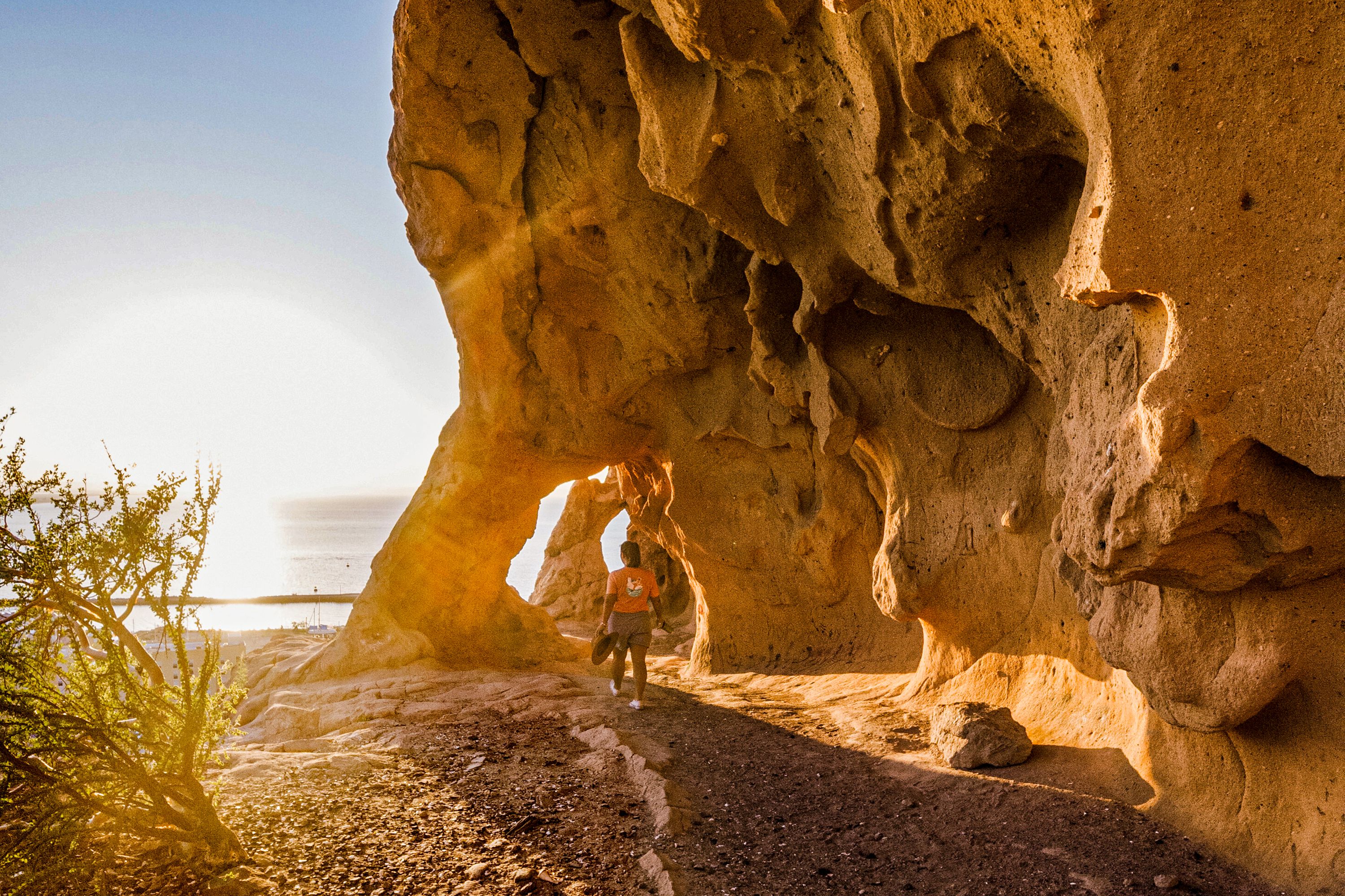 With her back to the camera a woman walks along a stone and dirt path next to a cliff and towards the sea and the sun low on the horizon.