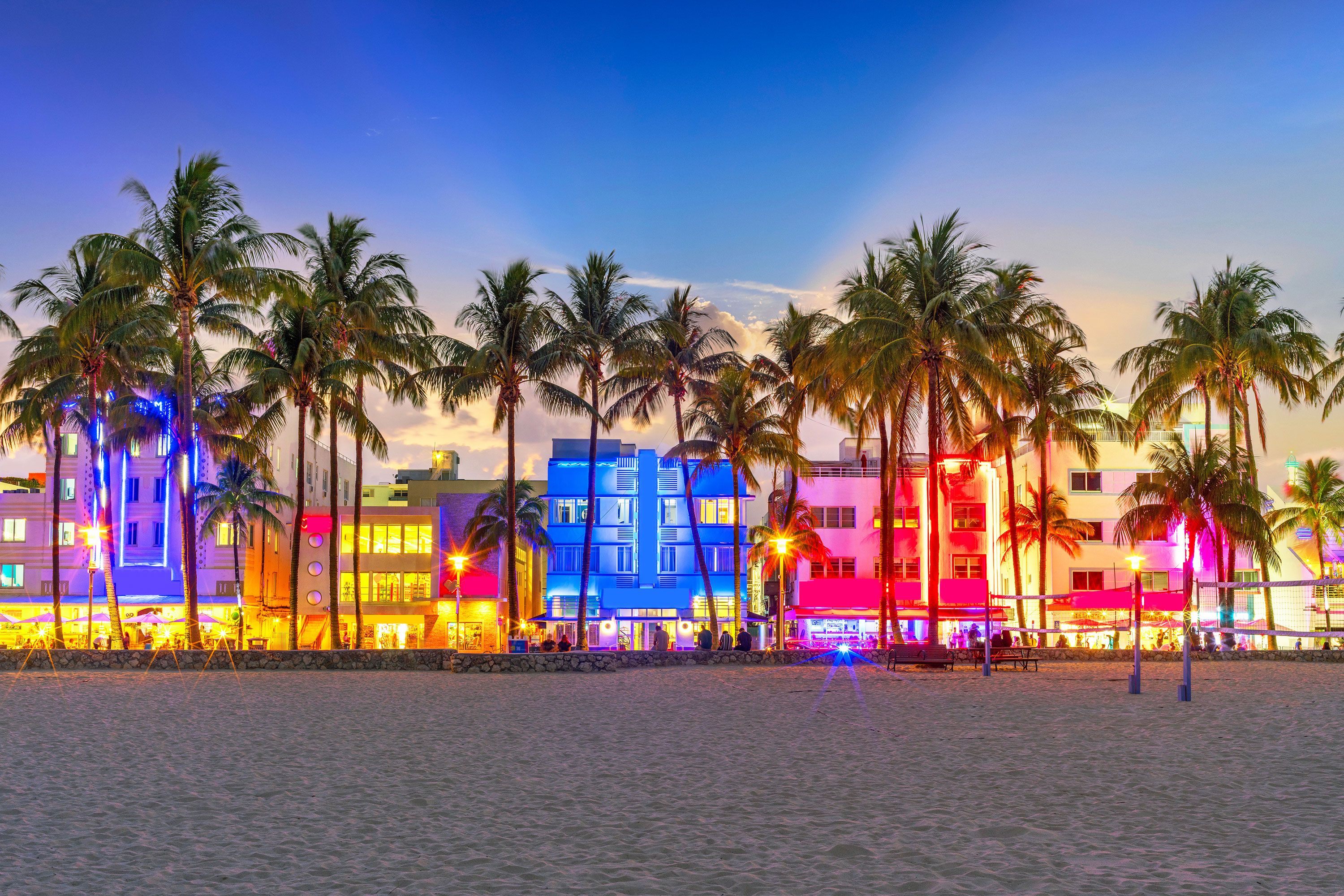 Colorful lights illuminate the buildings on Ocean Drive at night in Miami with palm trees and the beach in the front.