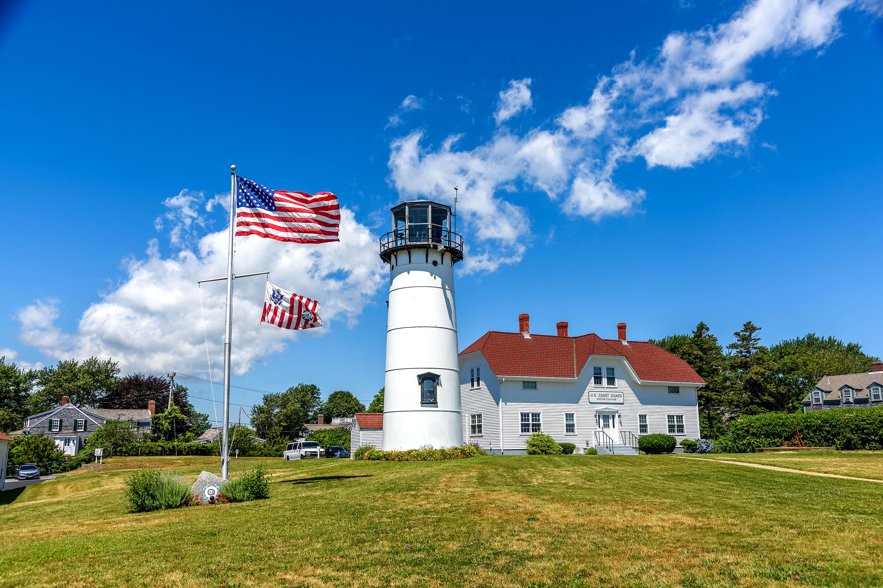 An American flag waves next to a large lighthouse in a grassy area.