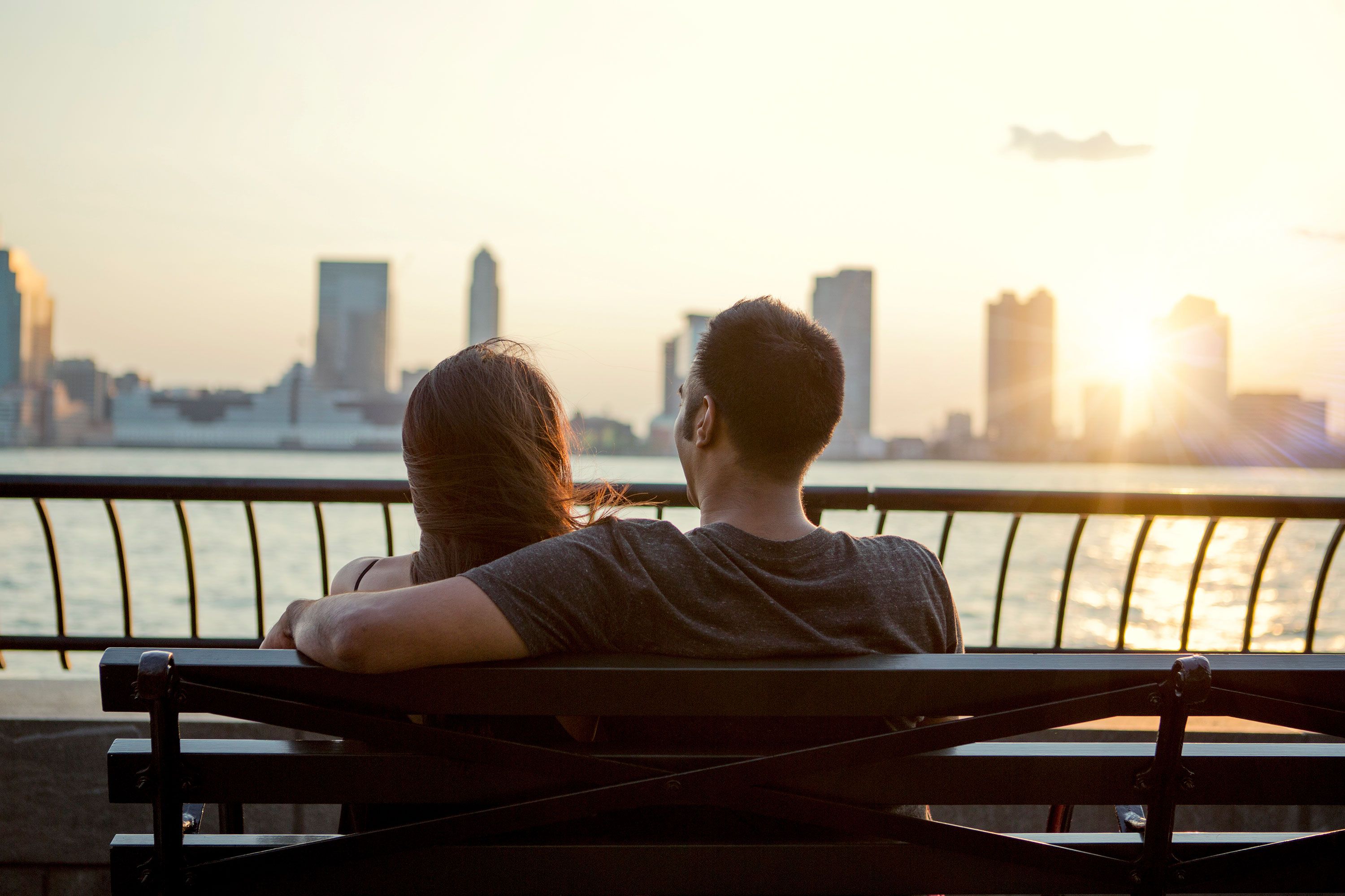 Rear view of two people sitting side by side on a park bench, looking over an urban river with skyscrapers on the far bank under a cloudy sky.