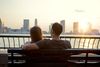 Rear view of two people sitting side by side on a park bench, looking over an urban river with skyscrapers on the far bank under a cloudy sky.