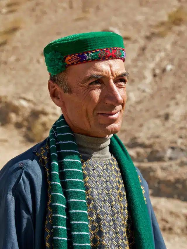 A man wearing a bright green and red embroidered hat and striped scarf squints into the sunlight