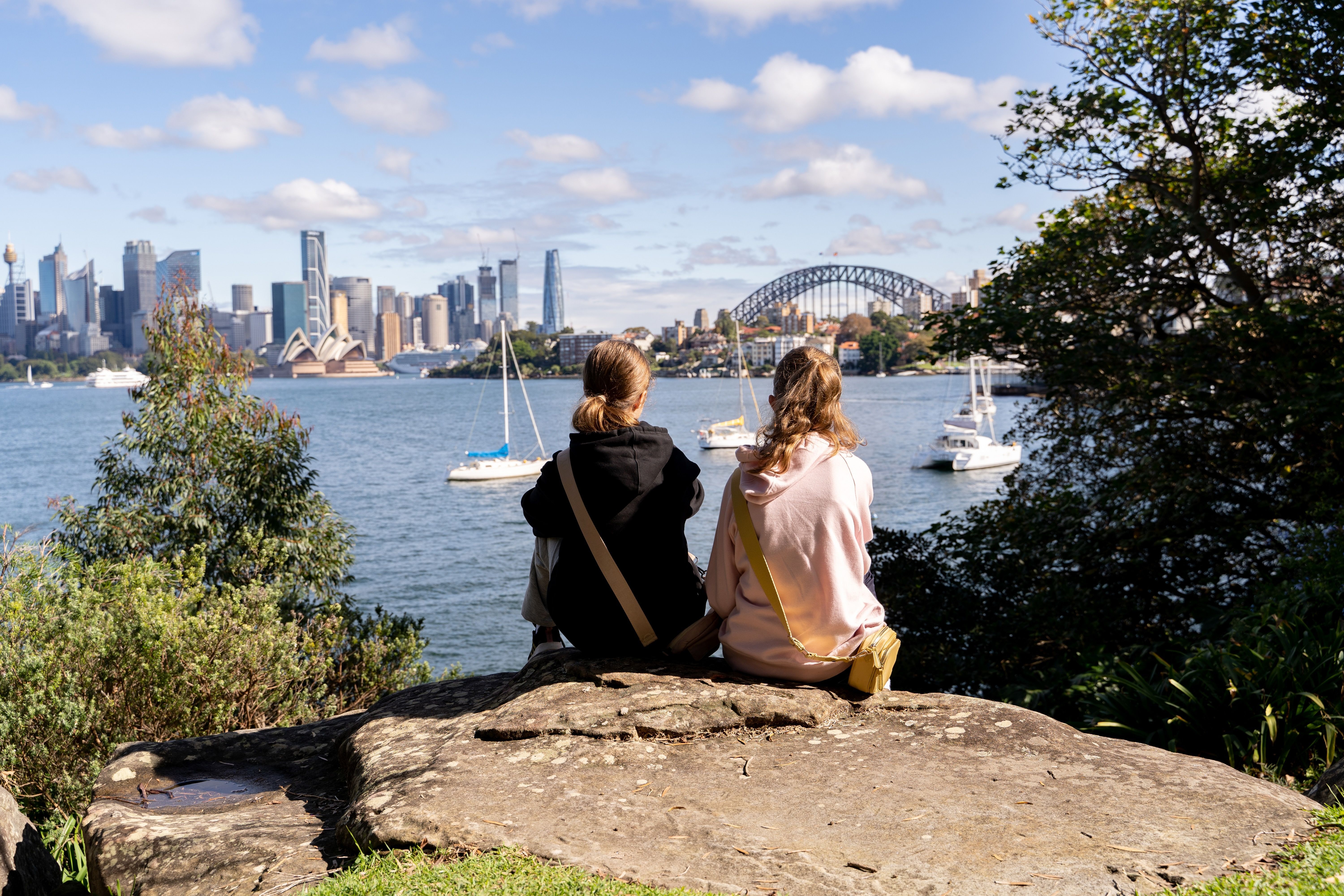 Two people seen from behind sit on a rock at the edge of the harbour looking out to the boats, city skyline and the Sydney Harbour Bridge in Australia