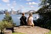 Two people seen from behind sit on a rock at the edge of the harbour looking out to the boats, city skyline and the Sydney Harbour Bridge in Australia
