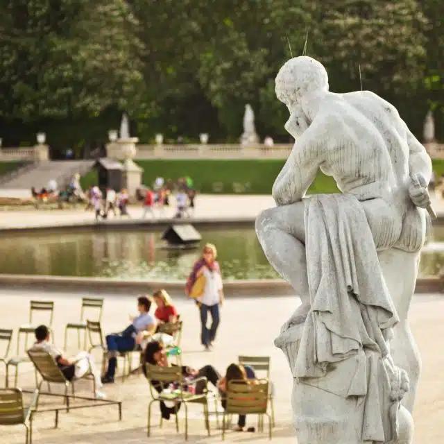 A classical sculpture of a man in Jardin de Luxembourg, Paris, with an ornamental lake seen behind and tourists seated and walking