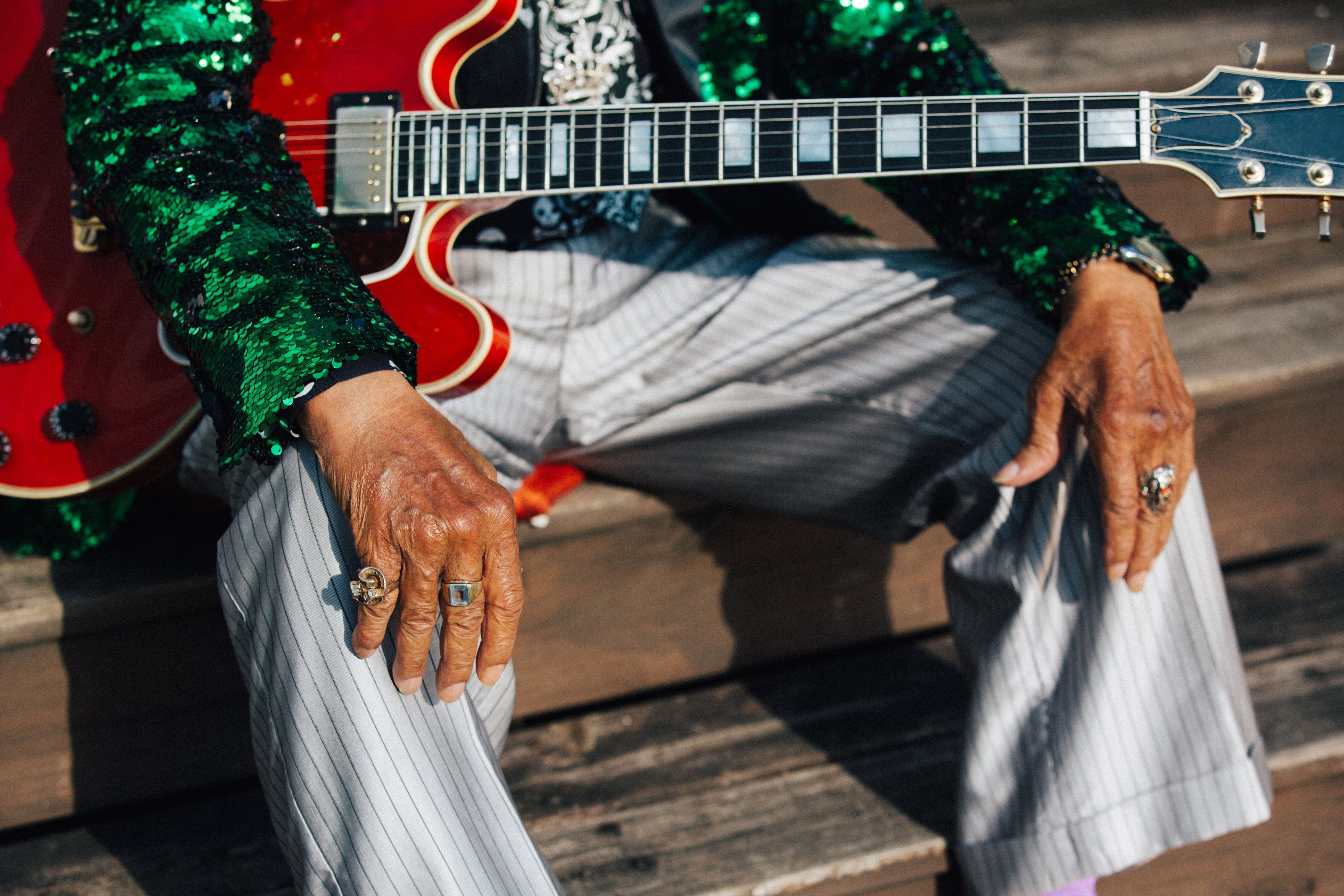 A close-up of a seated man wearing a green sequin jacket holding a red guitar