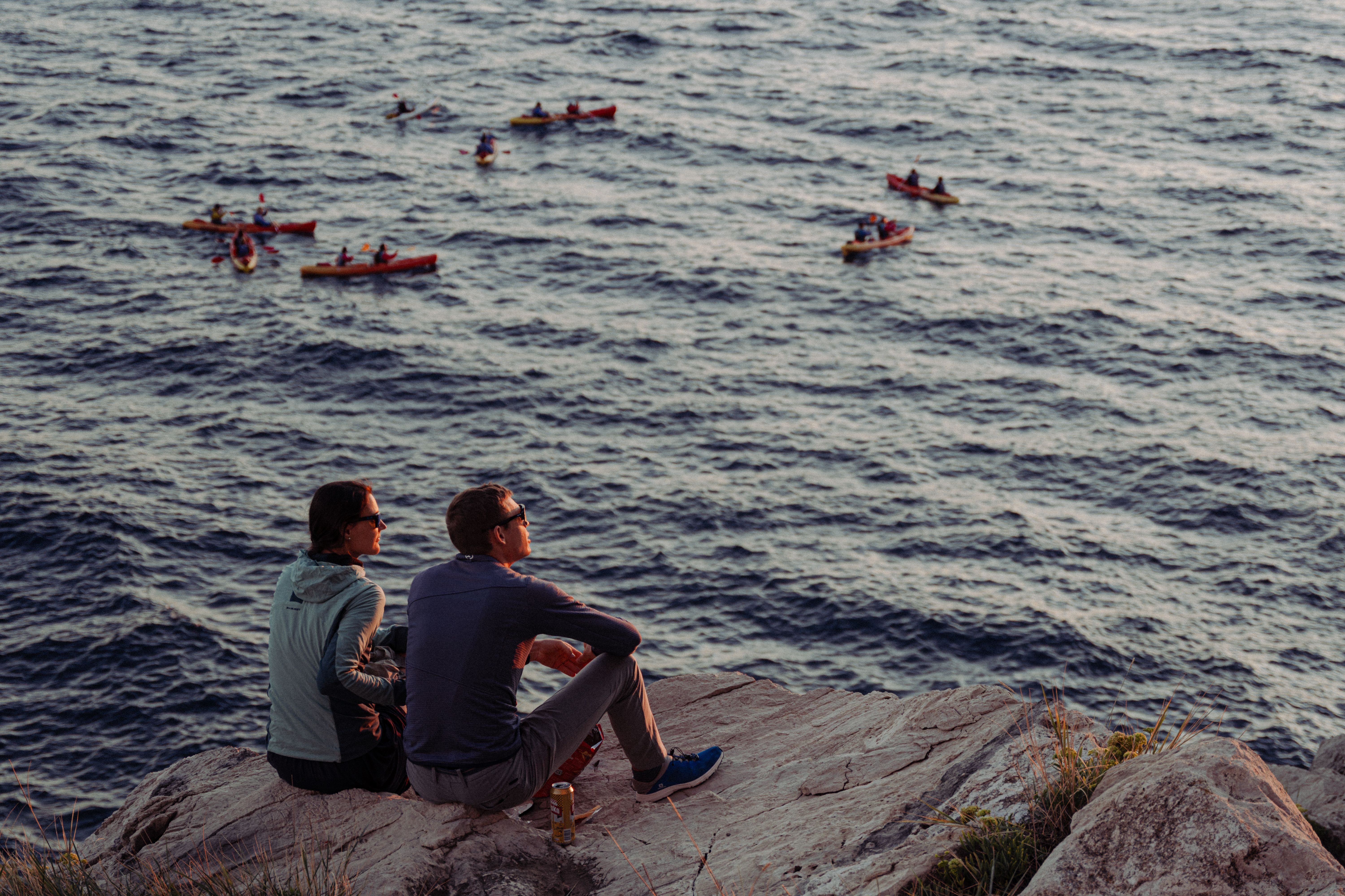 Couple sitting on a rocky cliff enjoying the view of kayakers on the Croatian coast during sunset.