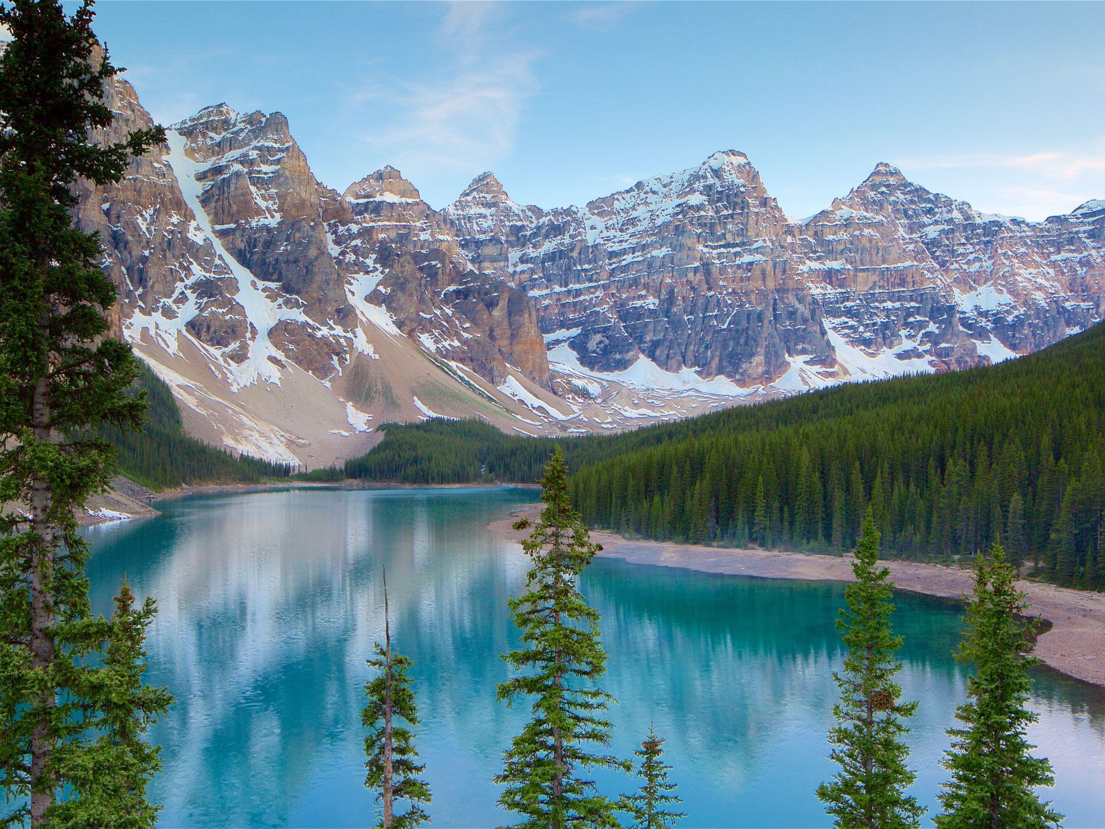 Large snowcapped mountains are seen before a vibrant blue lake with tall green trees in the foreground