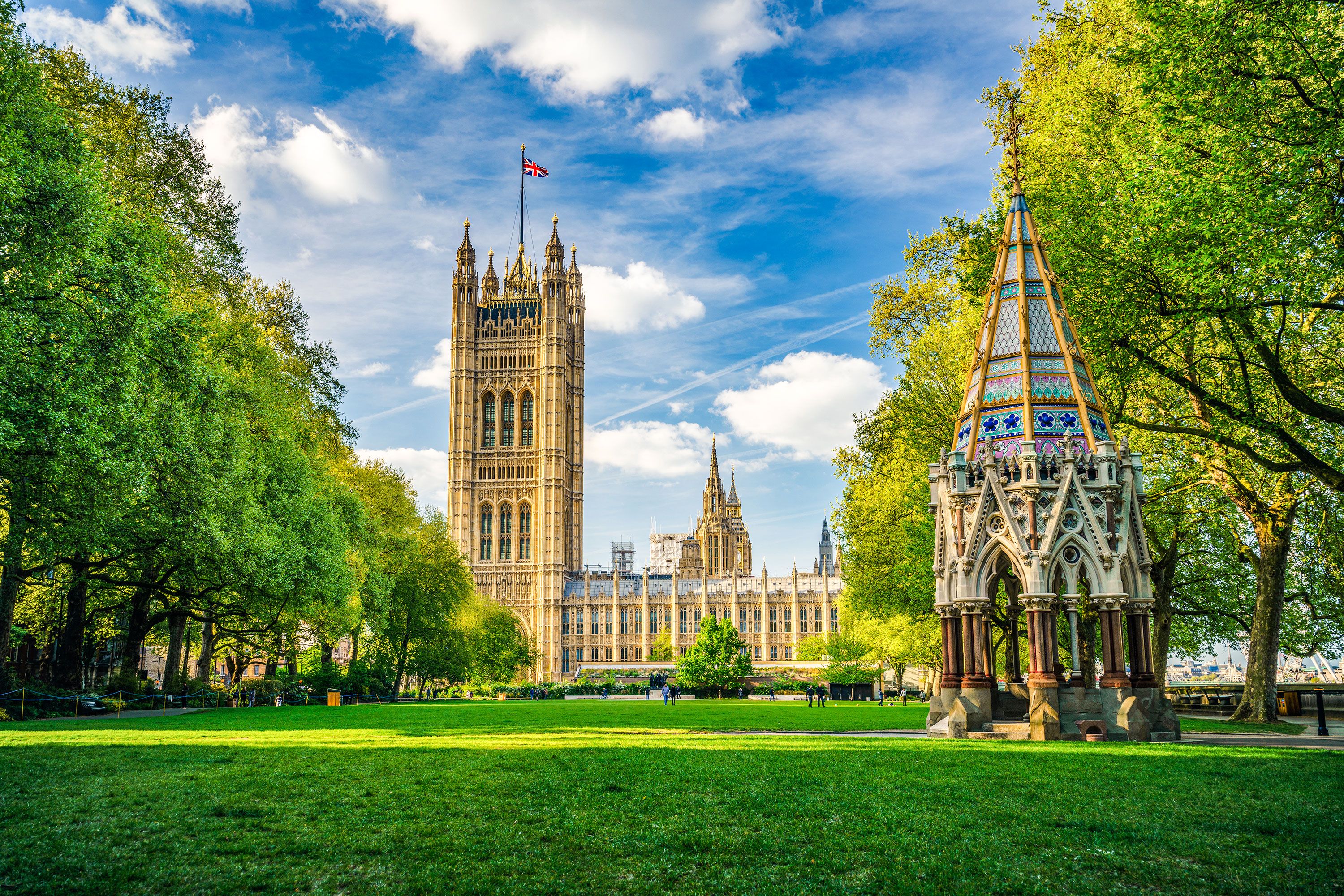 Victorian gothic monument on grassy lawn with tall tower and historic building behind.