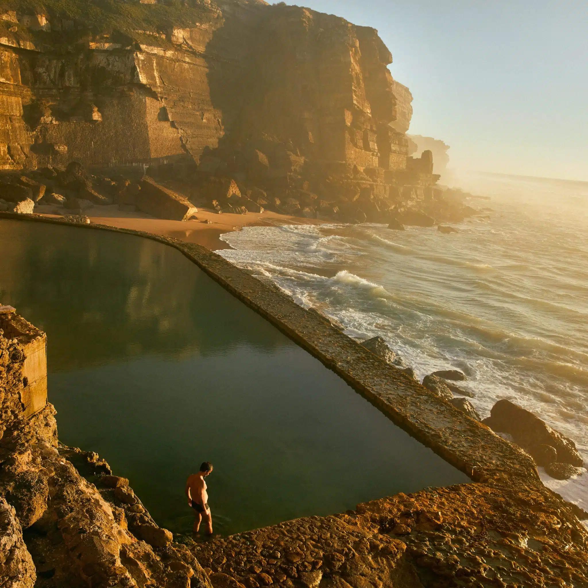 A swimmer stands at the edge of the seawater pool at Azenhas do Mar at sunset, which waves are just reaching from below