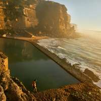 A swimmer stands at the edge of the seawater pool at Azenhas do Mar at sunset, which waves are just reaching from below