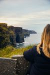 A person with long blond hair is seen from behind looking out onto the rugged Cliffs of Moher and the Atlantic Ocean on the West Coast of Ireland