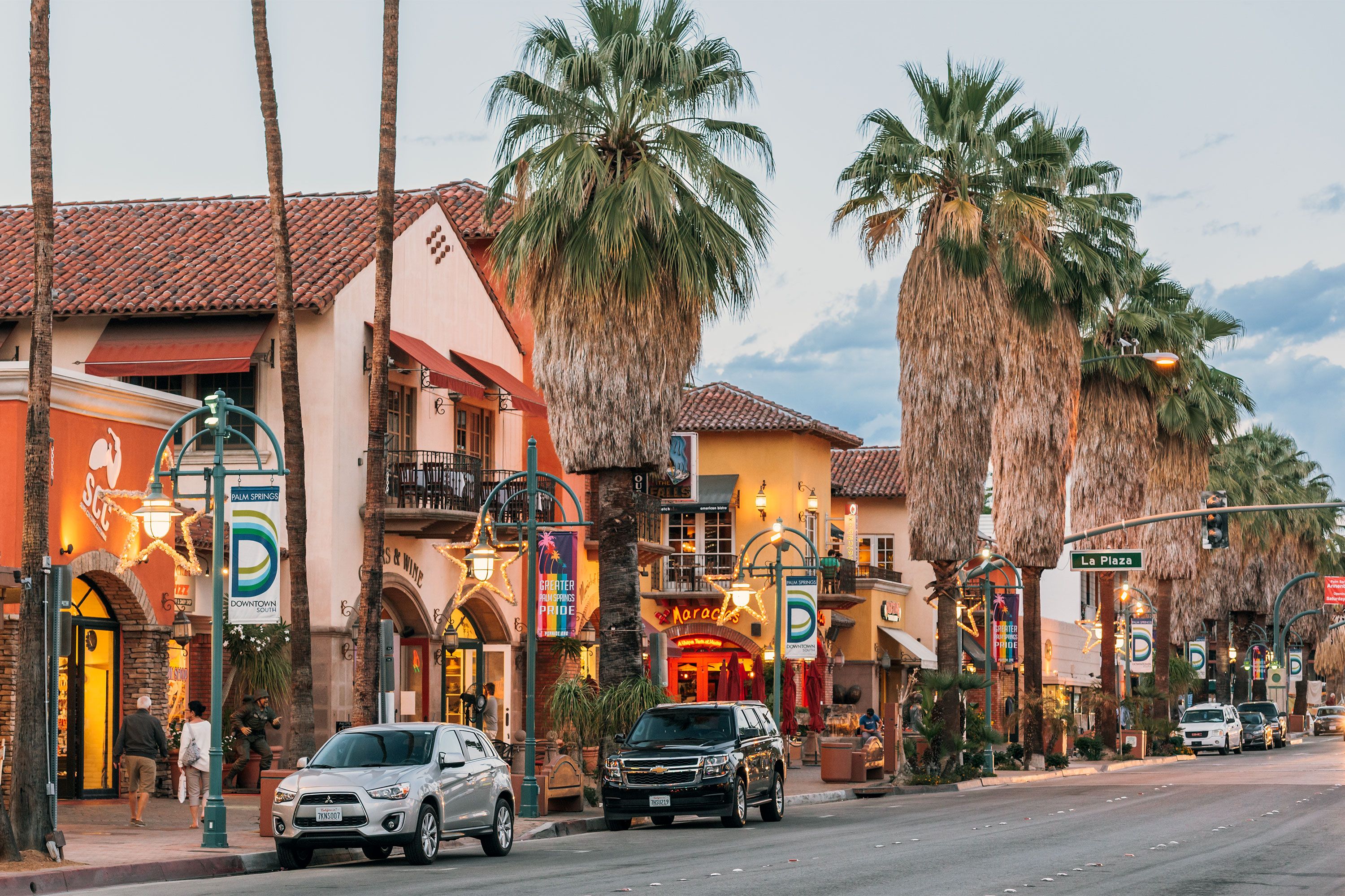 A street view of downtown Palm Springs, California, showing palm trees, Spanish-style buildings with tile roofs, colorful shops, and cars parked along the road at sunset.