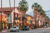 A street view of downtown Palm Springs, California, showing palm trees, Spanish-style buildings with tile roofs, colorful shops, and cars parked along the road at sunset.