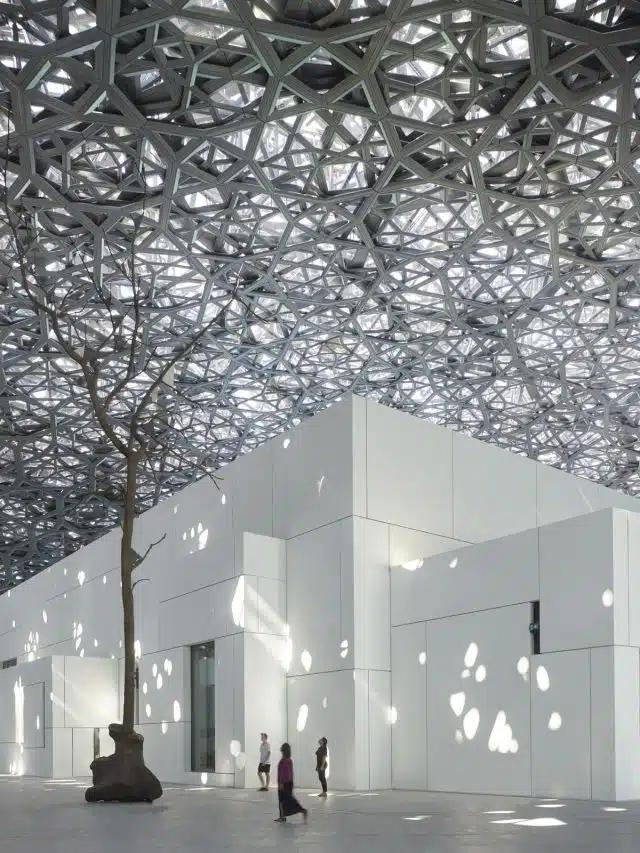 Light pours through a metal mesh roof onto a white cuboid building in the Louvre Abu Dhabi