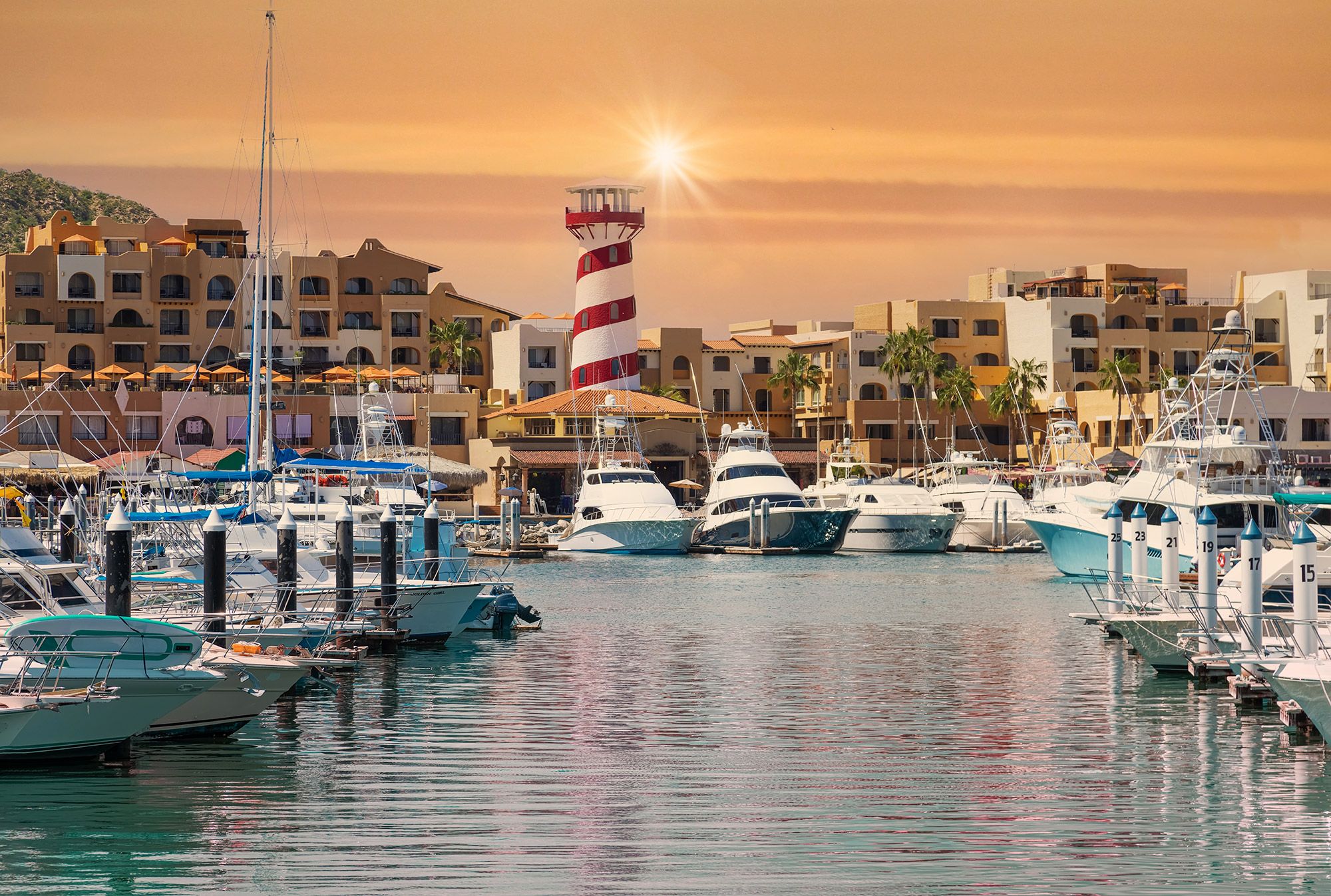 Boats docked at the marina in Cabo San Lucas with a red and white spiral lighthouse in the distance.
