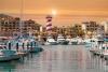 Boats docked at the marina in Cabo San Lucas with a red and white spiral lighthouse in the distance.