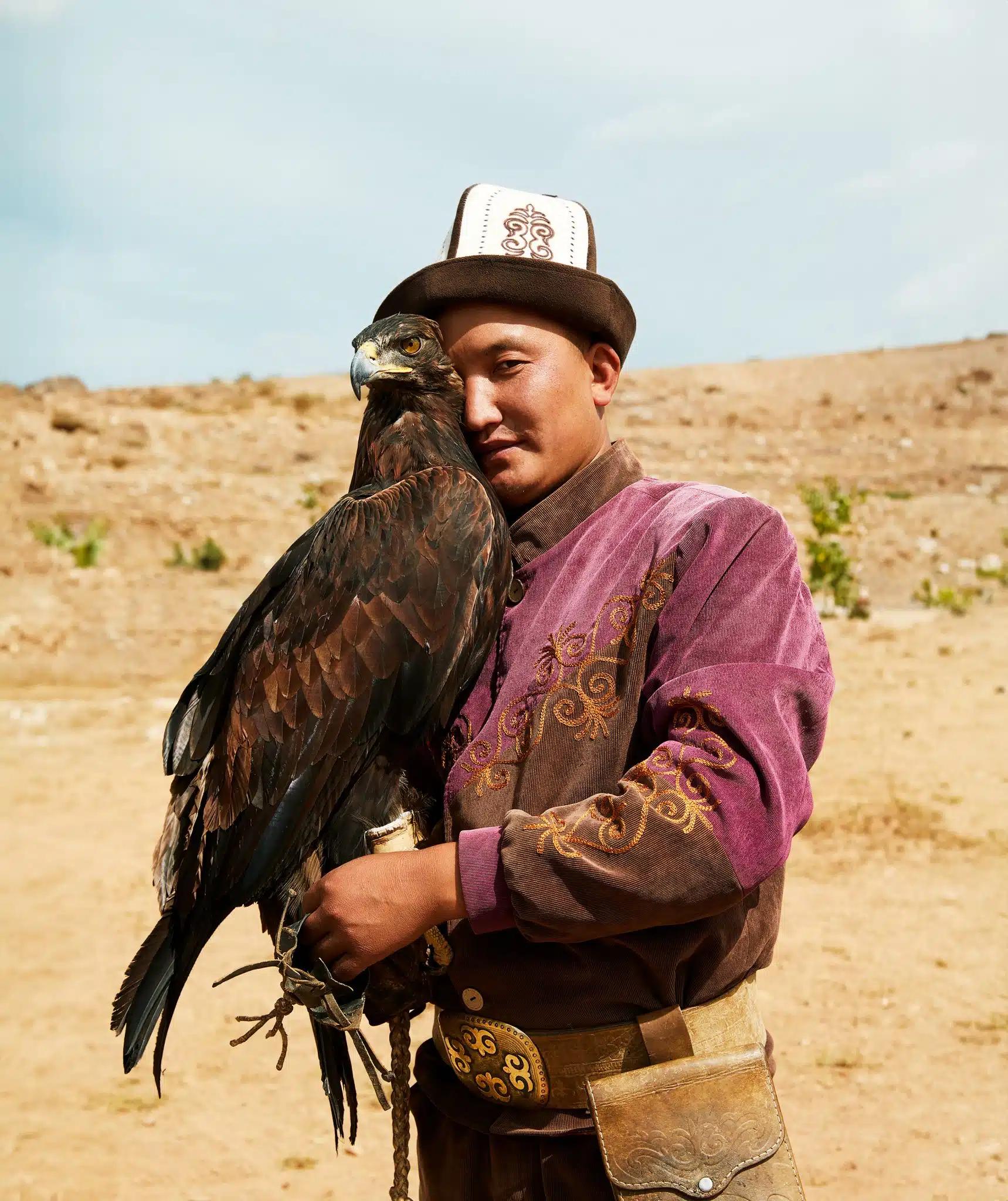 A man wearing an embroidered purple jacket and a hat with a large eagle perched on his hand; both look into the camera
