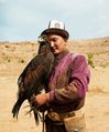 A man wearing an embroidered purple jacket and a hat with a large eagle perched on his hand; both look into the camera