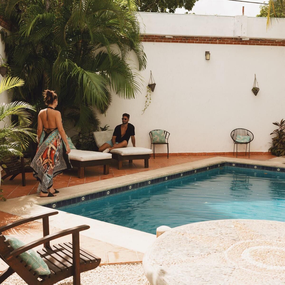 A woman in a flowing halterneck sundress walks towards a man sitting on a sun lounger by a swimming pool