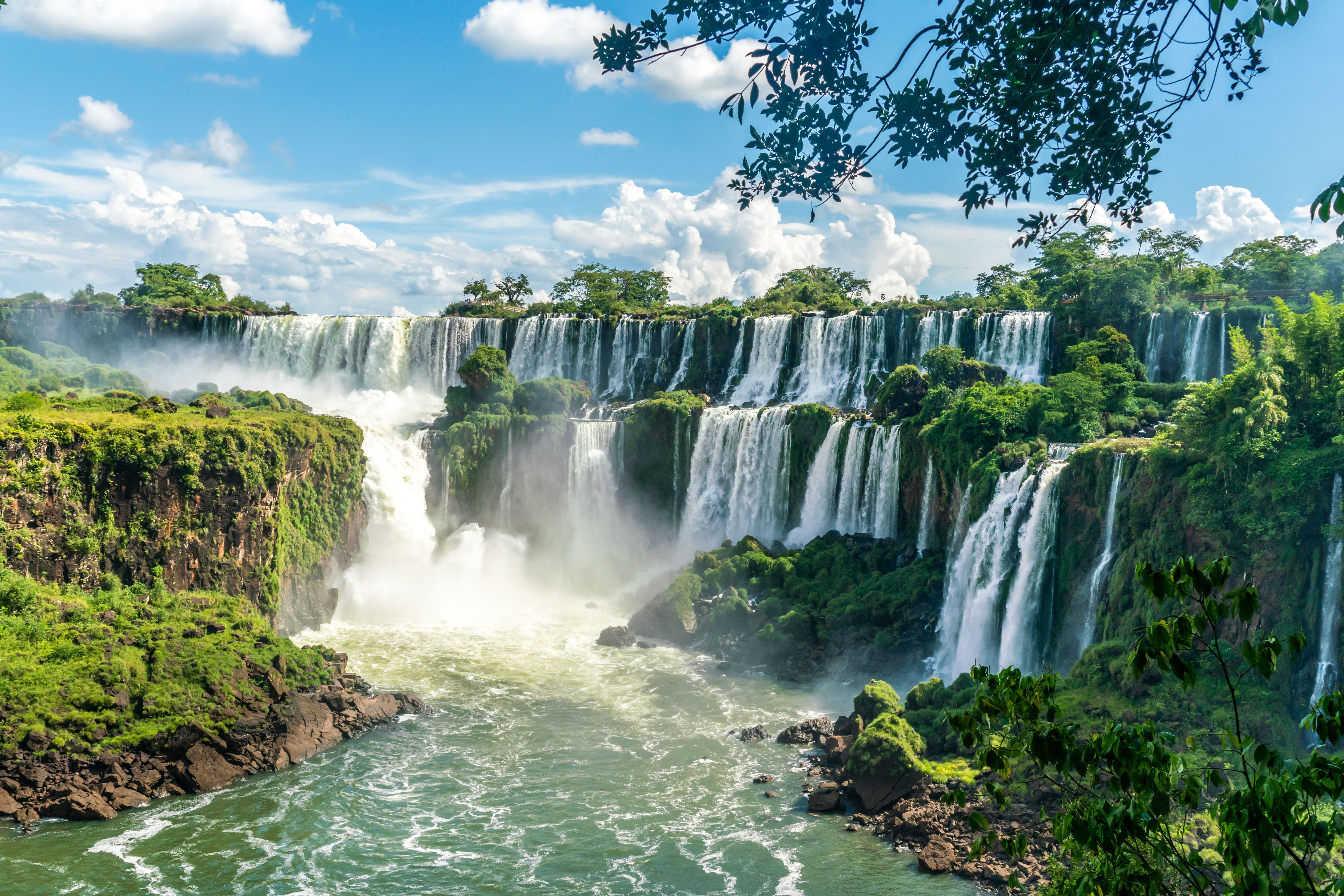 A large waterfall is seen falling over mossy rocks before a bright blue sky