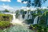 A large waterfall is seen falling over mossy rocks before a bright blue sky