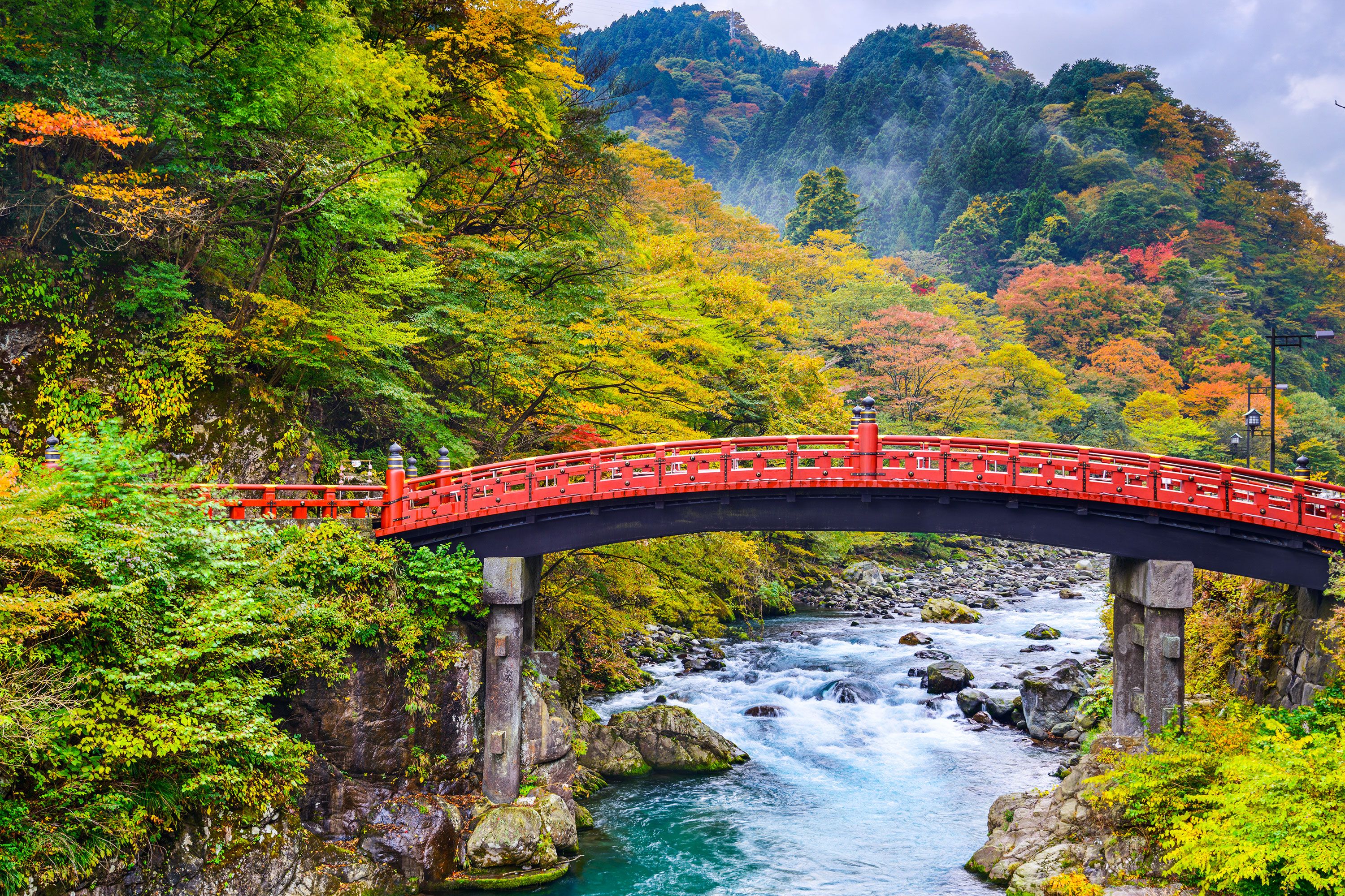 The iconic red Shinkyo Bridge arching over a green-toned river in Nikko, Japan, with trees lining both riverbanks.
