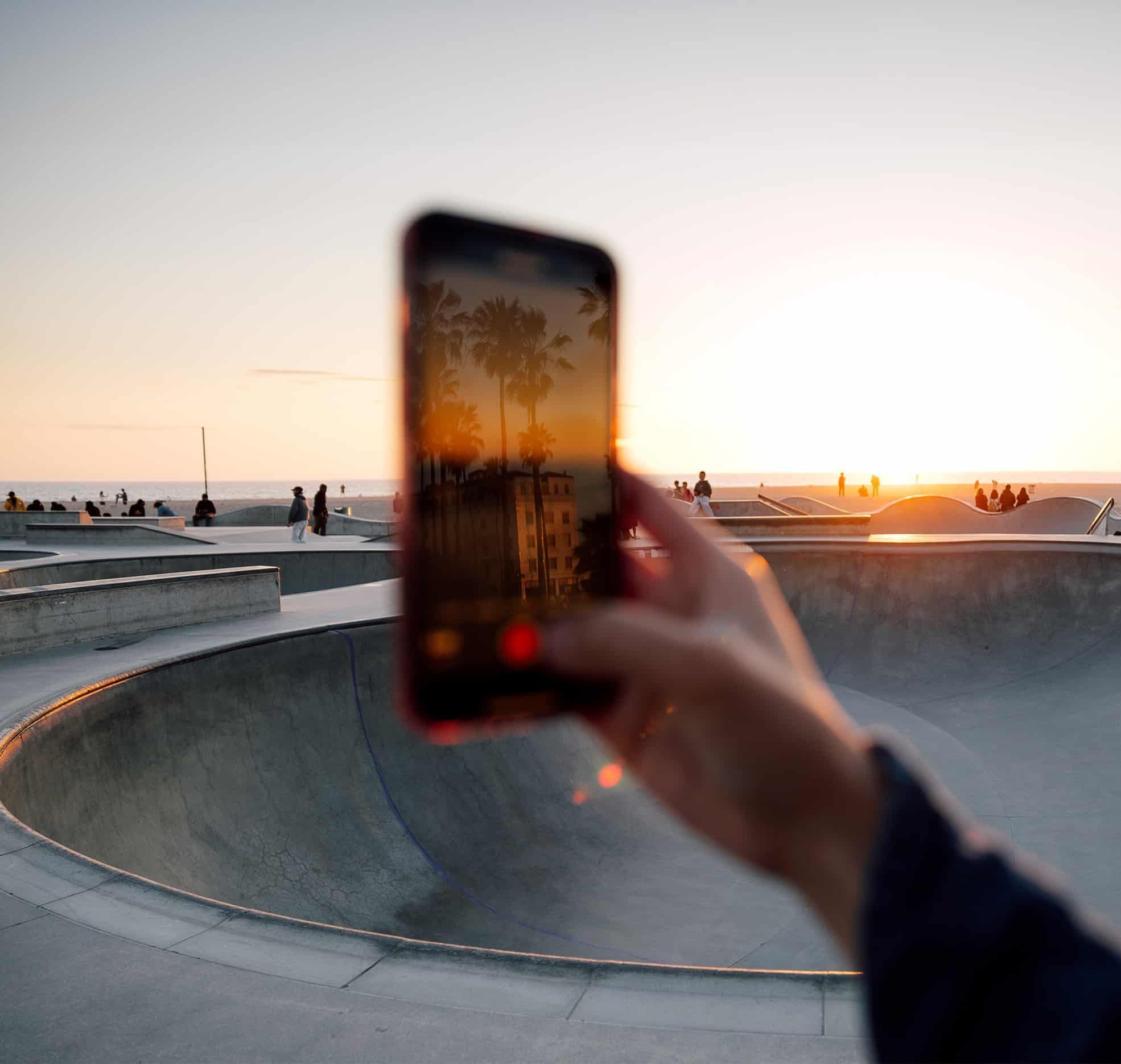 A person holds up a smartphone to take a photo of a sunset at Venice Beach Skate Park in California