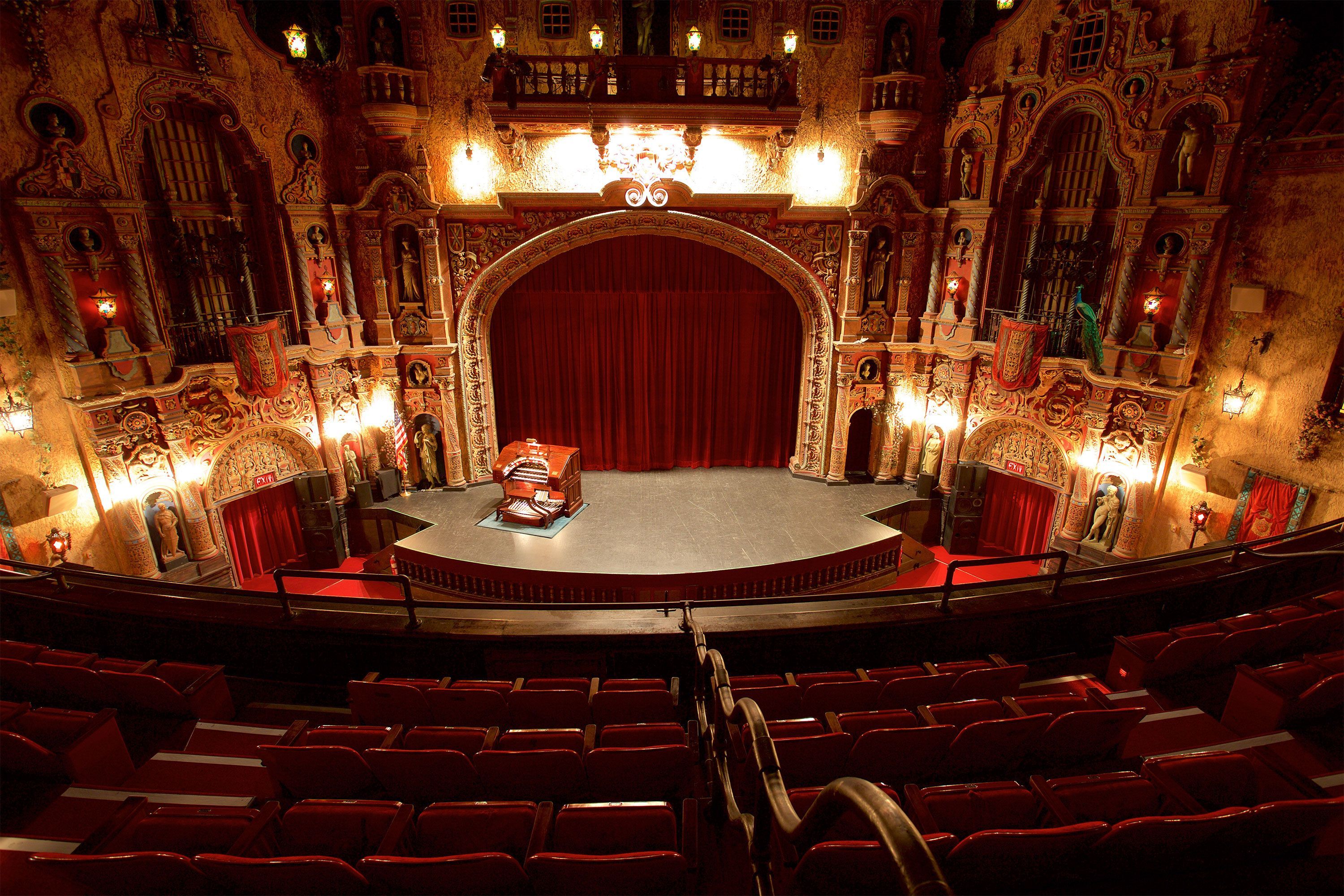 View from the balcony of an elaborately decorated theater interior, showing a red-curtained stage with a vintage organ and rows of red seats in the foreground.