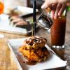 A plate of chicken and waffles, with syrup being poured over, and a bloody Mary