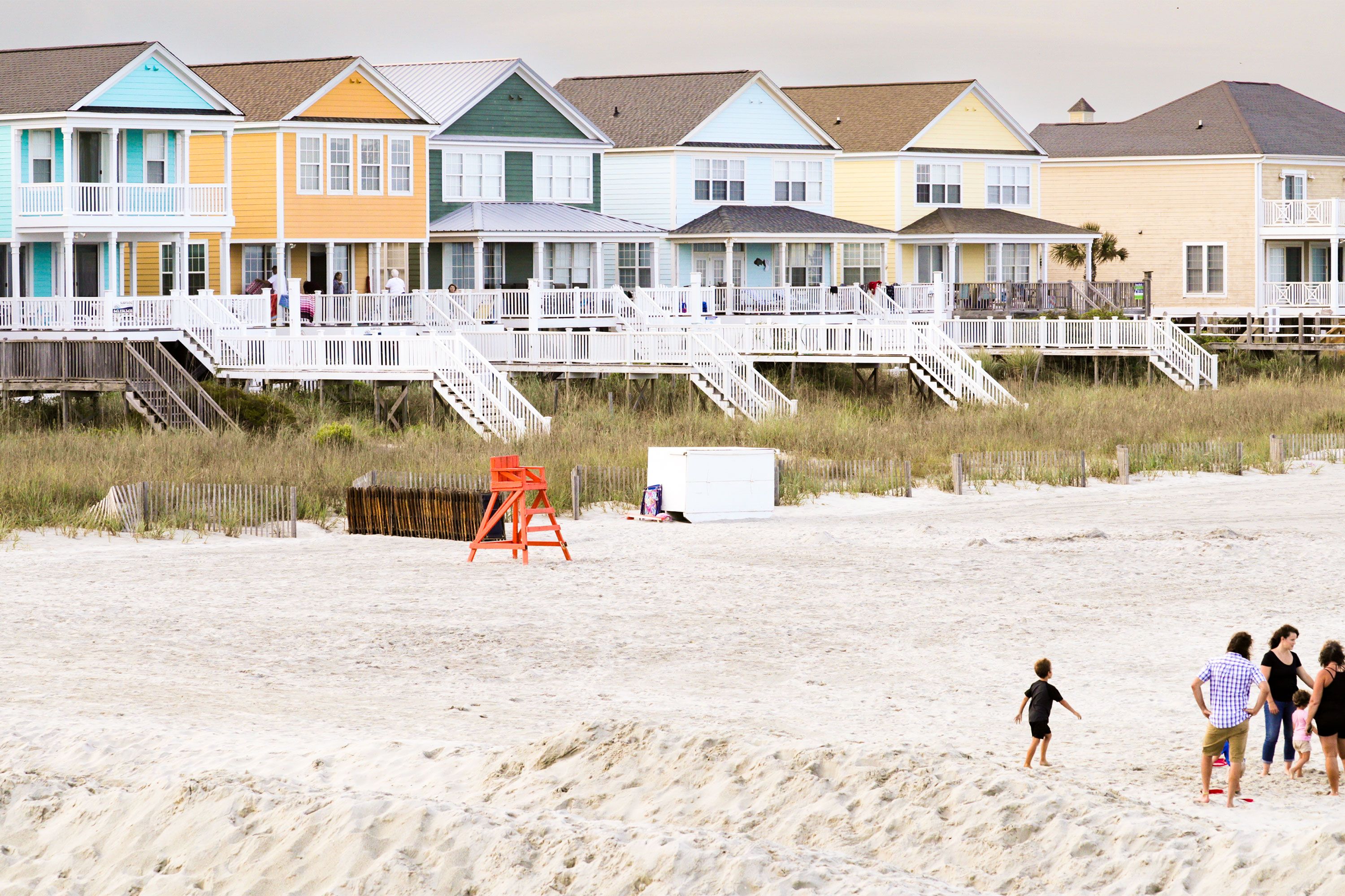 Colorful beach houses stand behind a sandy shoreline where people walk and play near the water.