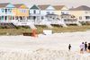 Colorful beach houses stand behind a sandy shoreline where people walk and play near the water.