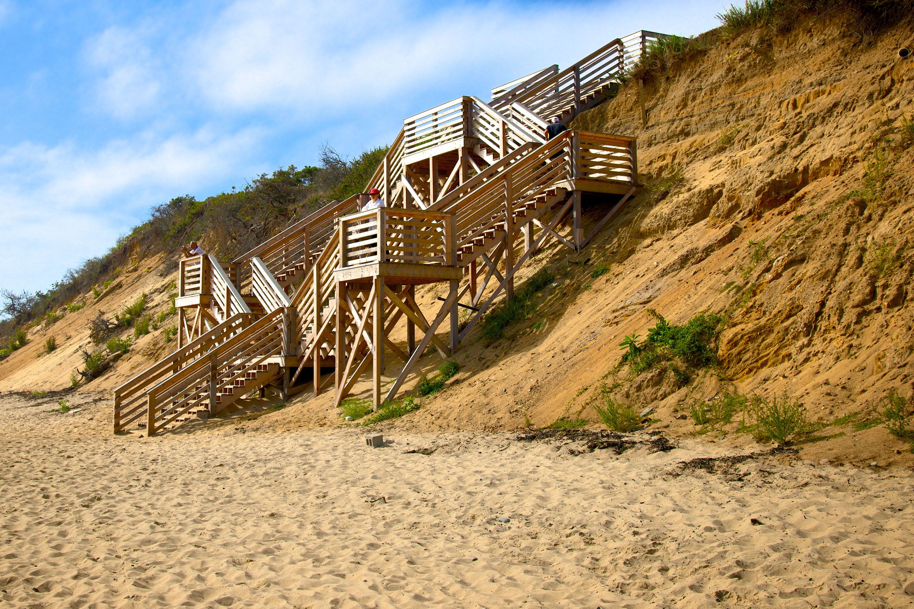 Wooden stairways built into a tall sandy dune provide access to the beach, with a few people standing on the platforms under a partly cloudy sky.