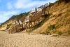 Wooden stairways built into a tall sandy dune provide access to the beach, with a few people standing on the platforms under a partly cloudy sky.
