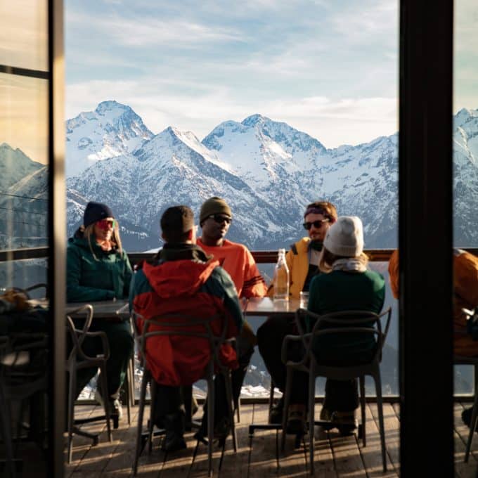 A group of young people sit out on the terrace of a bar with the snow-covered French Alps in the background