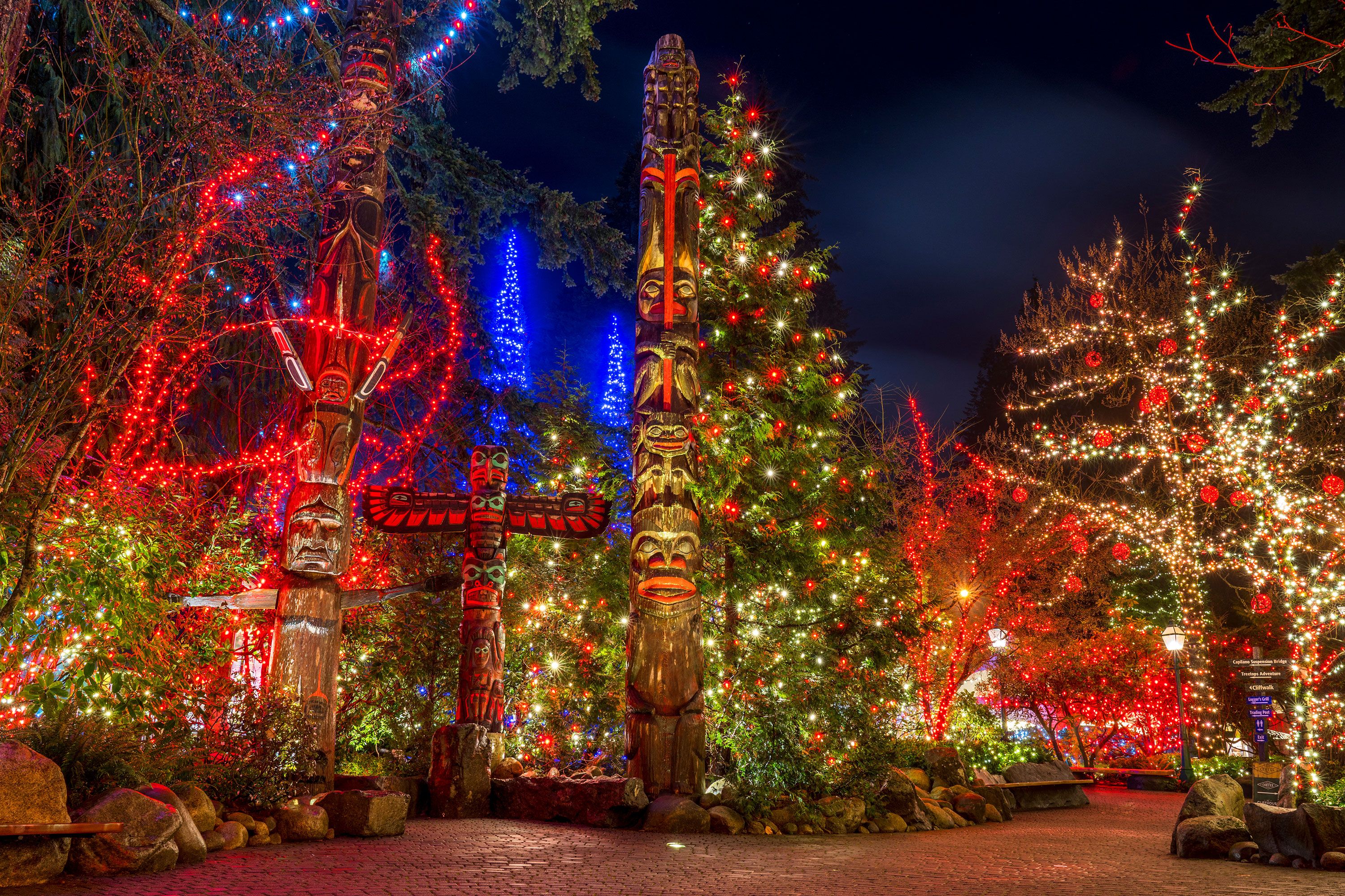 Totem poles at night illuminated by colorful Christmas lights in the background.