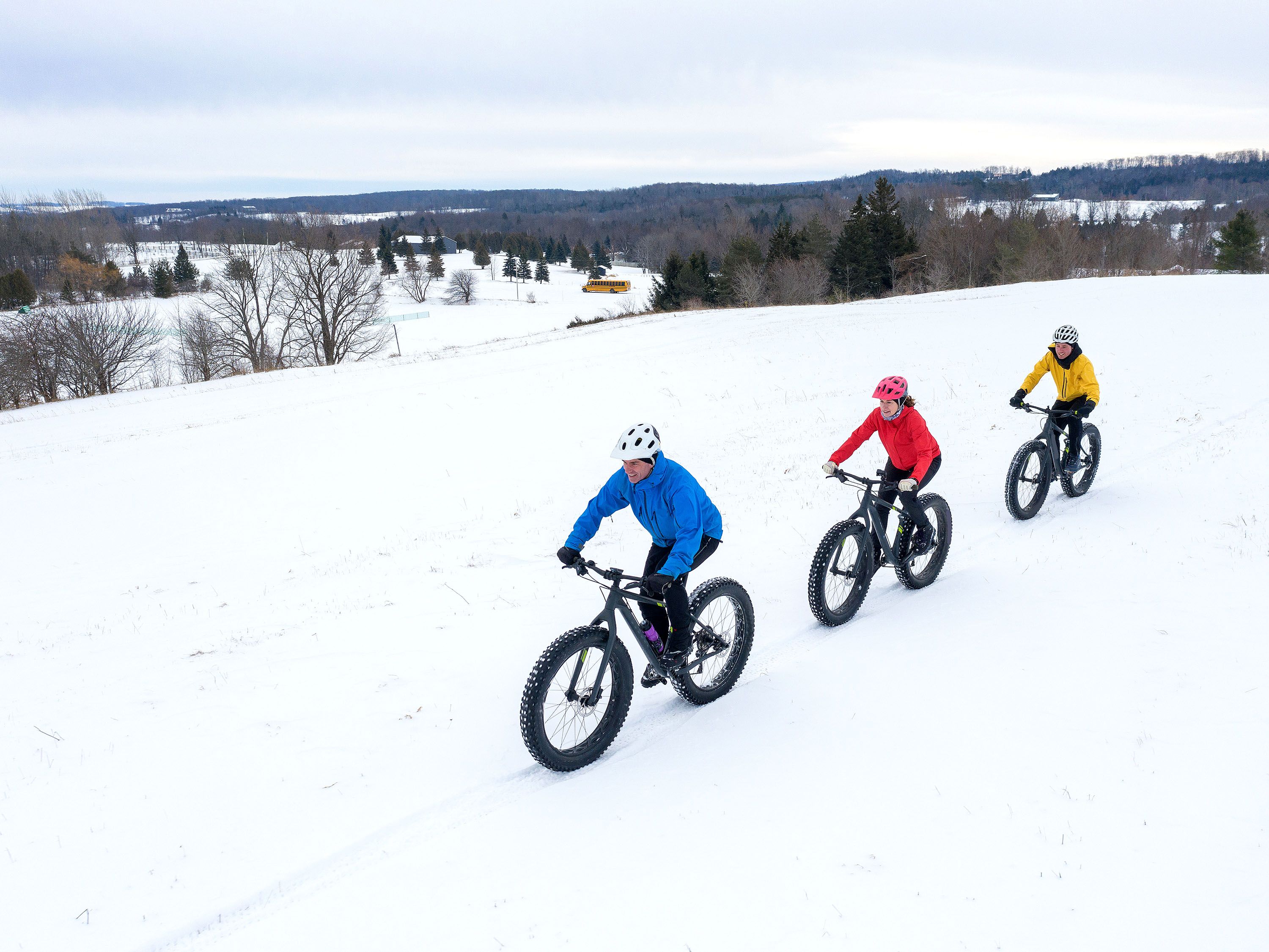 Three bicyclists in blue, red and yellow jackets ride in the snow.