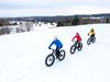 Three bicyclists in blue, red and yellow jackets ride in the snow.