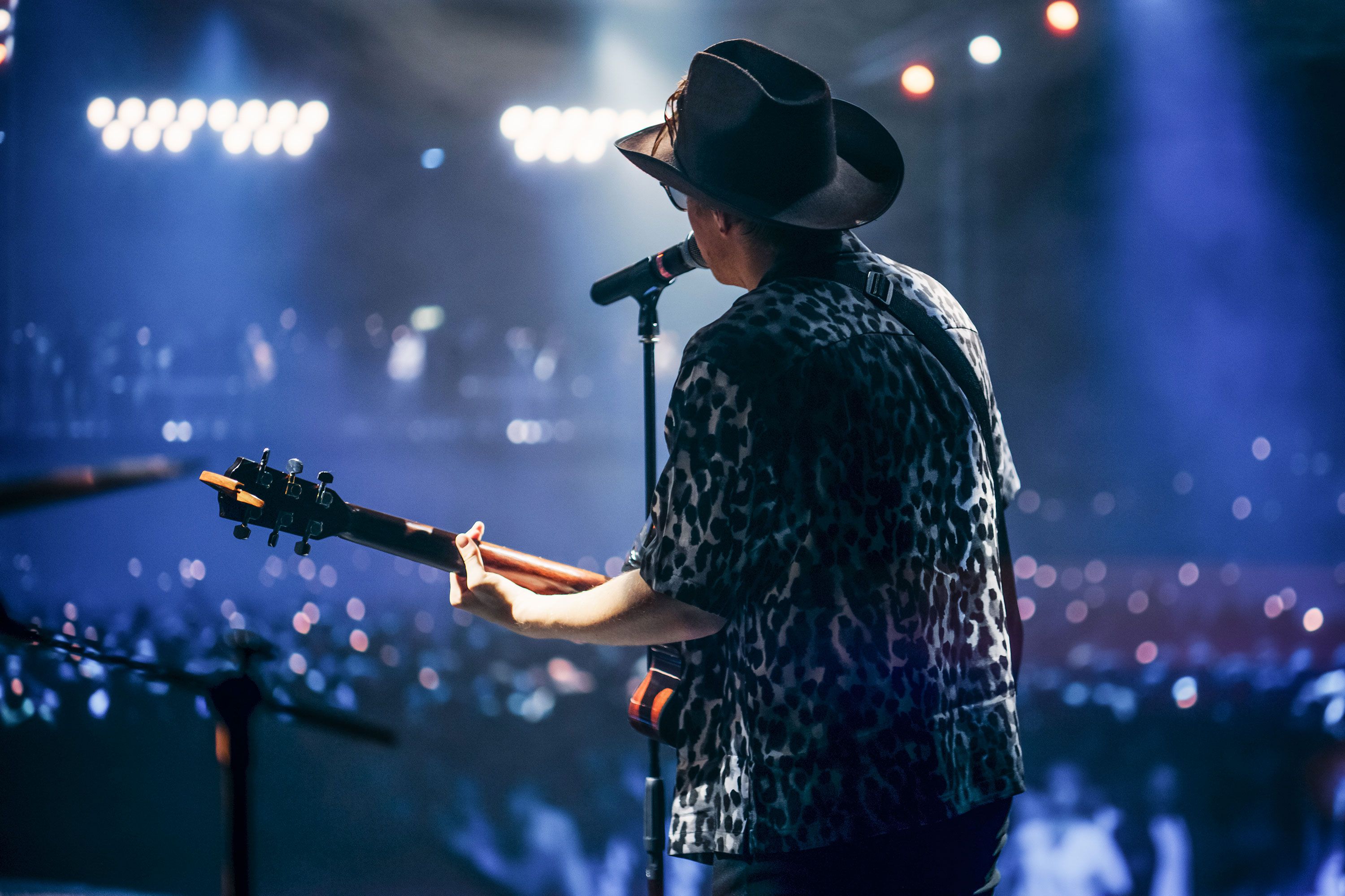 Perspectice from behind on stage of a musician with guitar and cowboy hat performing for crowd.