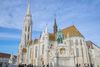 An exterior view of Matthias Church in Budapest showcasing the church’s Gothic spires, detailed façade, and distinctive multicolored glazed-tile roof under a bright sky.
