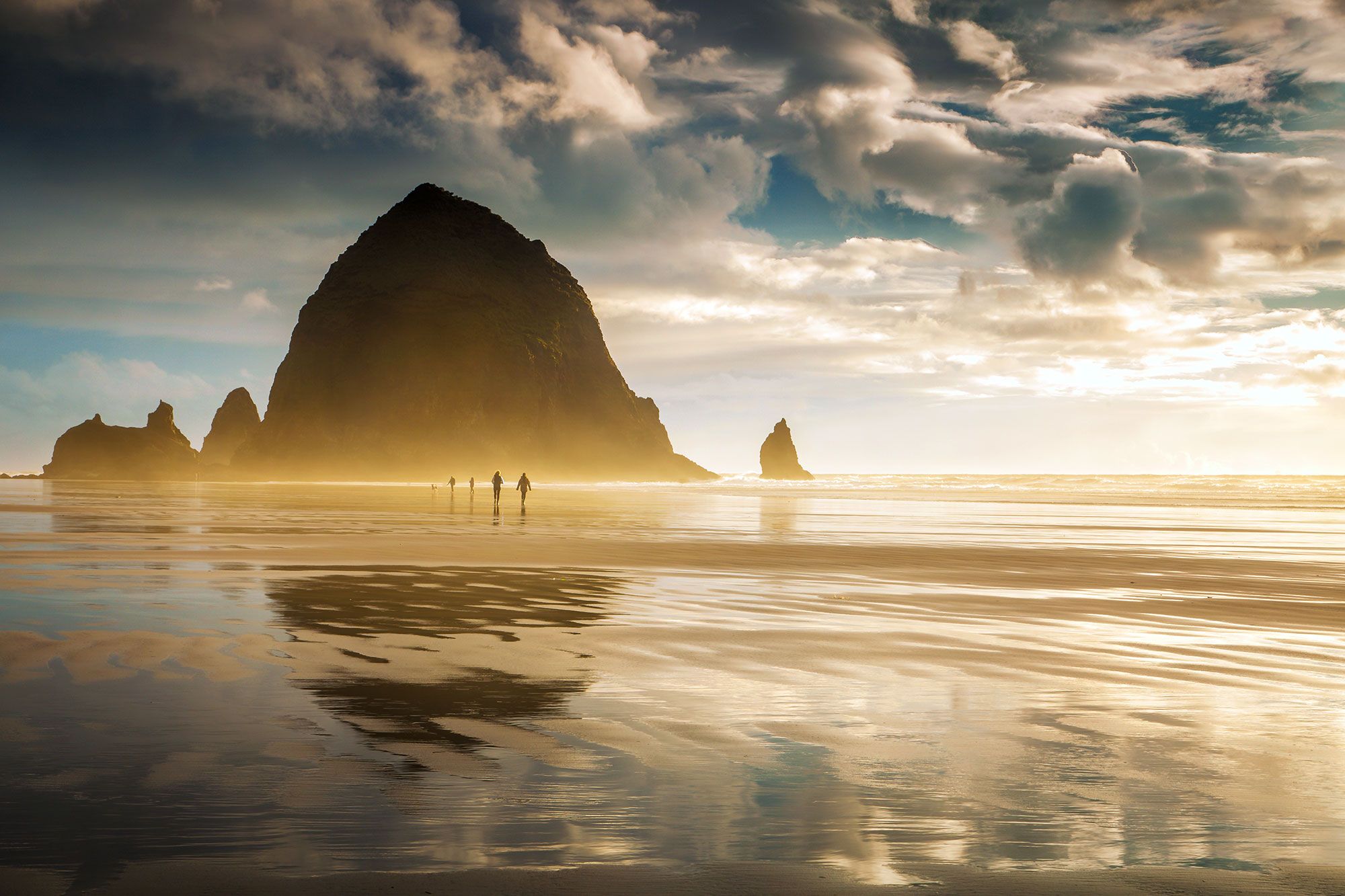 People walking on the beach in front of a massive rock.