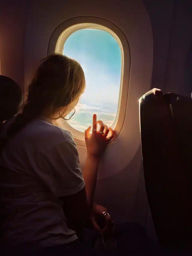 A woman looks out the window of an airplane to blue skies and sunlit clouds